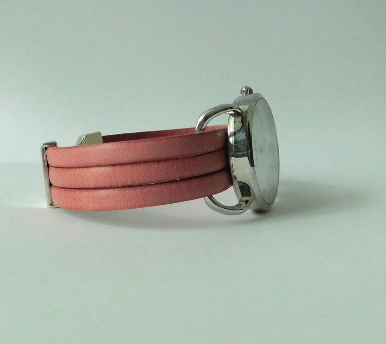 Puede incluir: Un reloj de pulsera con caja redonda de metal plateado y correa de varias vueltas hecha de cuero rosa liso. La correa consta de tres bandas distintas que se conectan a la caja del reloj con herrajes de metal plateado y una peque&ntilde;a corona.