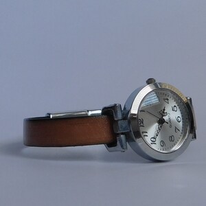 May include: A silver wristwatch with a brown leather band. The watch face is white with black numbers and hands. The watch face also says "QUARTZ".