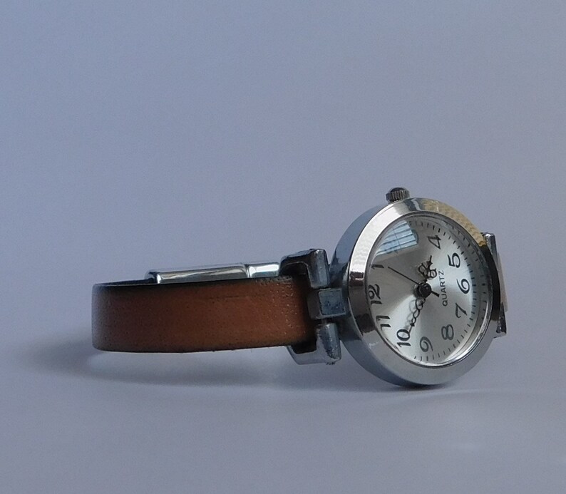 May include: A silver wristwatch with a brown leather band. The watch face is white with black numbers and hands. The watch face also says "QUARTZ".