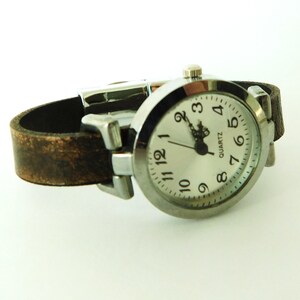 May include: A silver wristwatch with a brown leather band. The watch face is white with black numbers and hands. The watch face also says "QUARTZ".