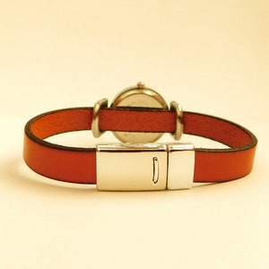 May include: A wristwatch with a round silver-colored face and a rust-colored leather band. The band is secured with a silver-colored magnetic clasp. The watch is designed to be worn on the wrist.