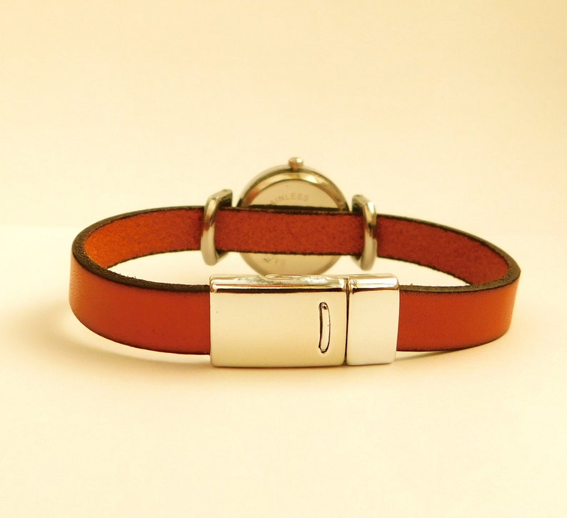 May include: A wristwatch with a round silver-colored face and a rust-colored leather band. The band is secured with a silver-colored magnetic clasp. The watch is designed to be worn on the wrist.