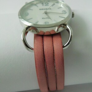 Puede incluir: Un reloj de pulsera con caja redonda de metal plateado y esfera blanca con el texto GENEVA ELITE. El reloj tiene una correa de varias hebras compuesta por tres bandas finas y lisas de piel sint&eacute;tica rosa empolvado unidas por bucles plateados.