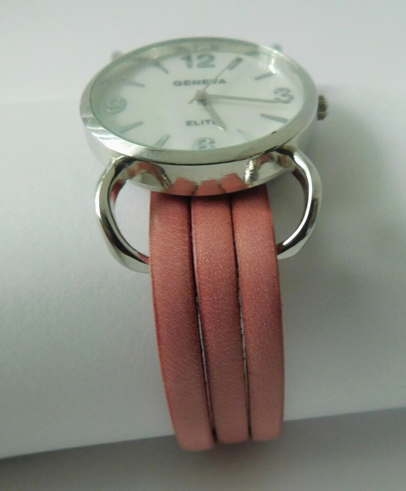 Puede incluir: Un reloj de pulsera con caja redonda de metal plateado y esfera blanca con el texto GENEVA ELITE. El reloj tiene una correa de varias hebras compuesta por tres bandas finas y lisas de piel sint&eacute;tica rosa empolvado unidas por bucles plateados.