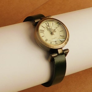 Op de afbeelding: Een gouden polshorloge met een witte wijzerplaat en zwarte Romeinse cijfers. Het horloge heeft een groene leren band.