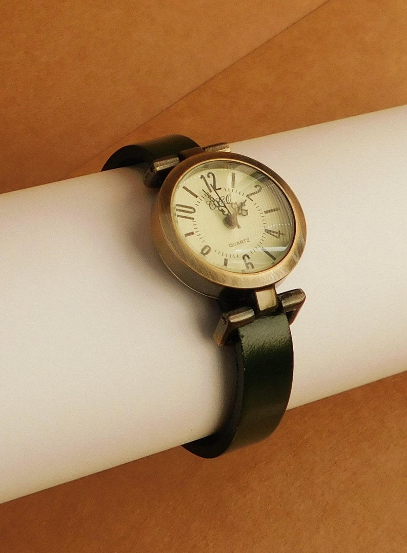 Op de afbeelding: Een gouden polshorloge met een witte wijzerplaat en zwarte Romeinse cijfers. Het horloge heeft een groene leren band.