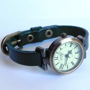 Op de afbeelding: Een groen leren polshorloge met een ronde, messingkleurige wijzerplaat en witte cijfers. Het horloge heeft een eenvoudig ontwerp met een klassieke uitstraling.
