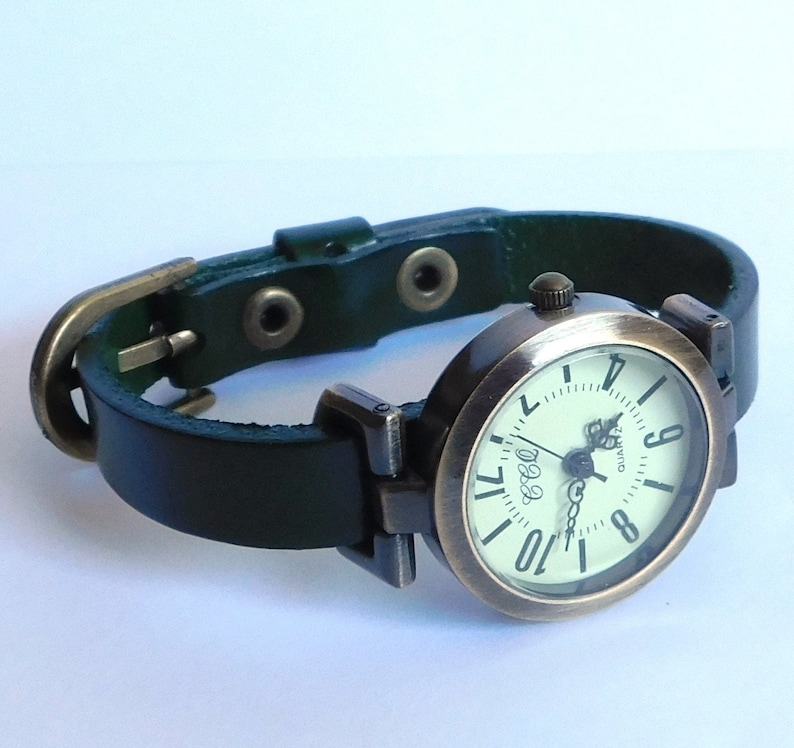 Op de afbeelding: Een groen leren polshorloge met een ronde, messingkleurige wijzerplaat en witte cijfers. Het horloge heeft een eenvoudig ontwerp met een klassieke uitstraling.