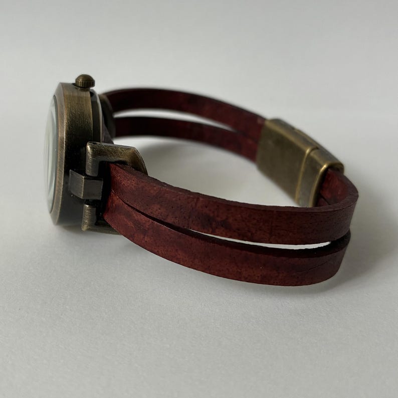 May include: A wristwatch featuring a round, dark bronze-toned metal case and a double-strand dark red leather strap. The watch includes a matching bronze-toned metal clasp connector, showcasing a textured, weathered finish on both the leather and metal components.