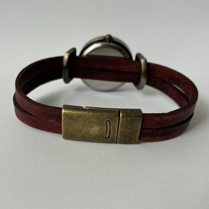 May include: A wristwatch featuring a dark brown leather strap that wraps around twice, secured by a rectangular antique brass-toned metal clasp. The watch case is circular with a metallic finish, connected to the leather band by brass-toned loops.