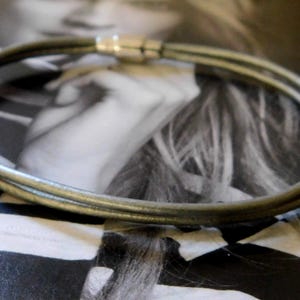 May include: A double-strand bracelet featuring thin, dark grey leather cords and a cylindrical silver-toned metal magnetic clasp. The bracelet is formed by two parallel loops of cord that meet at the metallic closure, creating a simple, circular design.