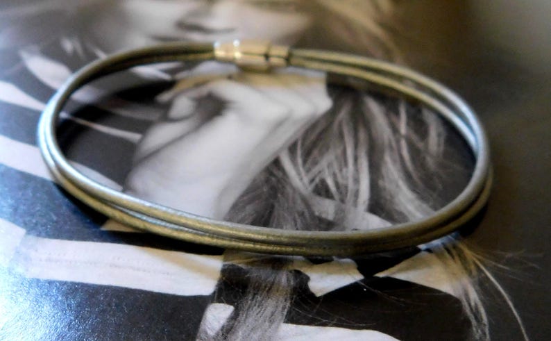 May include: A double-strand bracelet featuring thin, dark grey leather cords and a cylindrical silver-toned metal magnetic clasp. The bracelet is formed by two parallel loops of cord that meet at the metallic closure, creating a simple, circular design.