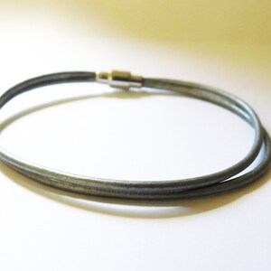 May include: A double-strand bracelet made of smooth, dark gray cord material. The two strands are joined together by a polished silver-toned cylindrical magnetic clasp that secures the ends of the cord, creating a simple and continuous circular loop.