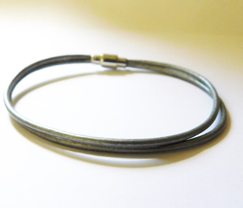 May include: A double-strand bracelet made of smooth, dark gray cord material. The two strands are joined together by a polished silver-toned cylindrical magnetic clasp that secures the ends of the cord, creating a simple and continuous circular loop.