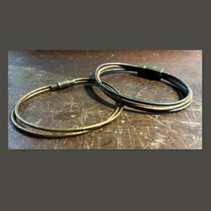 May include: Two multi-strand bracelets featuring thin, round leather cords in shades of brown and black. Each bracelet is secured with a cylindrical, antique-toned metal magnetic clasp that connects the multiple strands into a single loop.