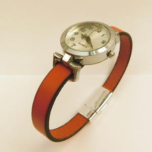 May include: A silver quartz watch with a round face and white dial, featuring black numerals and hands. The watch has a rust-colored leather band with a silver clasp. The watch band is approximately 0.5 inches wide.