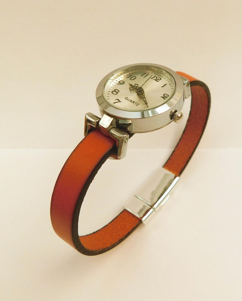 May include: A silver quartz watch with a round face and white dial, featuring black numerals and hands. The watch has a rust-colored leather band with a silver clasp. The watch band is approximately 0.5 inches wide.