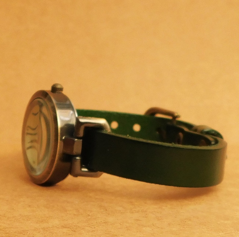 Op de afbeelding: Een zilveren polshorloge met een groene leren band. De wijzerplaat van het horloge heeft een uniek ontwerp met een helder glazen deksel.