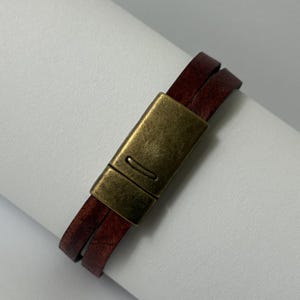 May include: A double-wrap bracelet featuring two strands of dark reddish-brown leather cord secured by a rectangular, antique-style brass magnetic clasp with a smooth, brushed metal finish and a small horizontal indentation on the front surface.