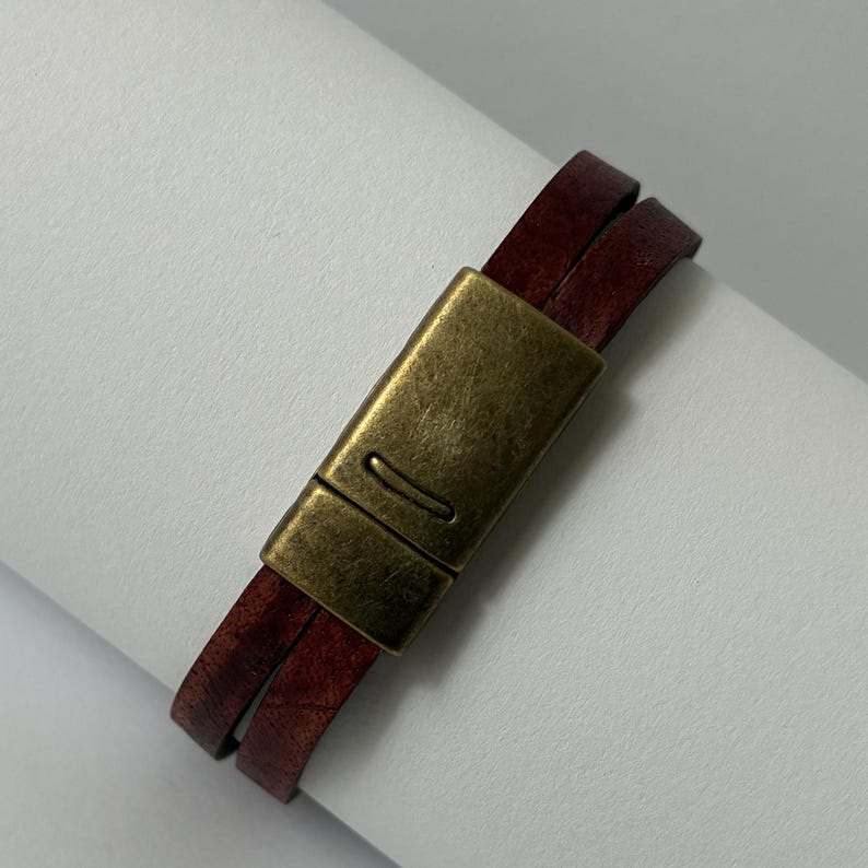 May include: A double-wrap bracelet featuring two strands of dark reddish-brown leather cord secured by a rectangular, antique-style brass magnetic clasp with a smooth, brushed metal finish and a small horizontal indentation on the front surface.