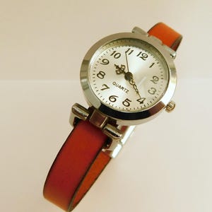 May include: A silver quartz watch with a white face and black numerals. The watch has a red-orange leather band. The watch face reads "QUARTZ". The watch band is approximately 0.5 inches wide.