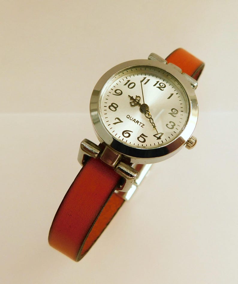 May include: A silver quartz watch with a white face and black numerals. The watch has a red-orange leather band. The watch face reads "QUARTZ". The watch band is approximately 0.5 inches wide.
