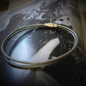 May include: A double-strand bracelet featuring a thin, metallic-toned cord with a smooth texture. The two strands are connected by a cylindrical gold-toned metal clasp mechanism that secures the circular shape of the jewelry piece.