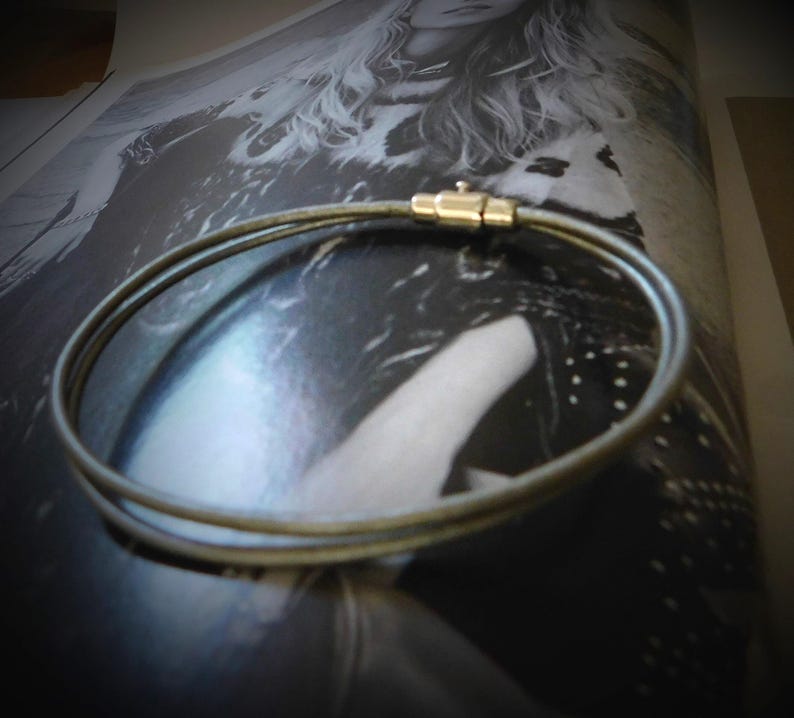 May include: A double-strand bracelet featuring a thin, metallic-toned cord with a smooth texture. The two strands are connected by a cylindrical gold-toned metal clasp mechanism that secures the circular shape of the jewelry piece.