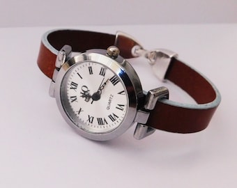 May include: A silver watch with a white face and black Roman numeral hour markers. The watch has a brown leather band.