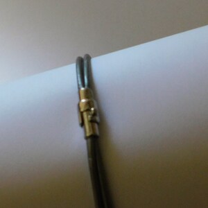 May include: A double-strand dark gray leather bracelet featuring a polished silver-tone metal cylindrical clasp. The two thin leather cords are secured together by the metallic fastener, creating a simple and clean design for the wrist.