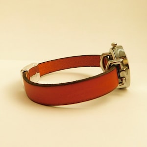 May include: A wristwatch with a red leather band and a silver-colored watch face. The band is secured with a silver-colored clasp. The watch face is round and has a silver-colored bezel.