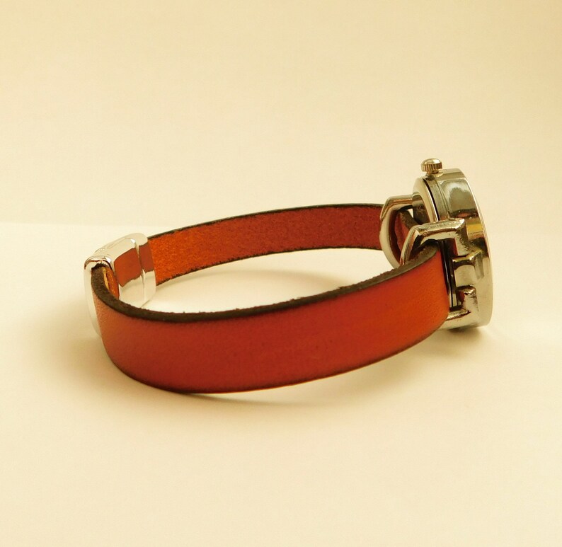 May include: A wristwatch with a red leather band and a silver-colored watch face. The band is secured with a silver-colored clasp. The watch face is round and has a silver-colored bezel.