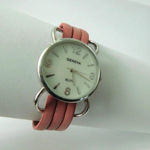 Puede incluir: Un reloj de pulsera Geneva Elite con caja redonda plateada, esfera blanca, manecillas plateadas y n&uacute;meros en las posiciones de las 3, 6, 9 y 12. El reloj est&aacute; sujeto a una correa de cuero rosa de varias hebras con bucles de metal plateado.