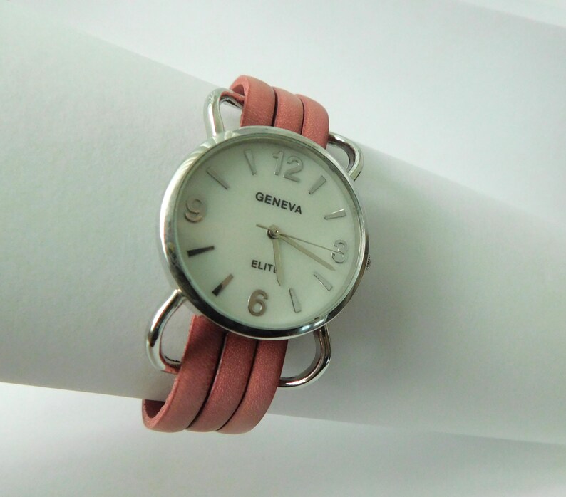 Puede incluir: Un reloj de pulsera Geneva Elite con caja redonda plateada, esfera blanca, manecillas plateadas y n&uacute;meros en las posiciones de las 3, 6, 9 y 12. El reloj est&aacute; sujeto a una correa de cuero rosa de varias hebras con bucles de metal plateado.