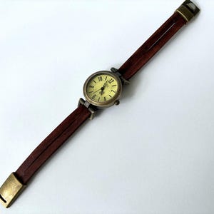 May include: A wristwatch with a round, antique brass-toned case and a pale yellow dial featuring black numerals. The watch band consists of two thin, dark brown leather straps connected by brass-toned metal hardware and a matching rectangular clasp.