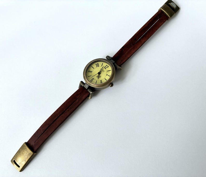 May include: A wristwatch with a round, antique brass-toned case and a pale yellow dial featuring black numerals. The watch band consists of two thin, dark brown leather straps connected by brass-toned metal hardware and a matching rectangular clasp.