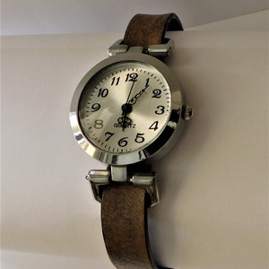 May include: A silver-toned wristwatch with a white face and black numbers. The watch has a brown leather band.