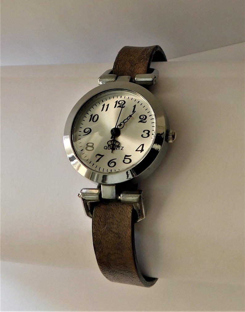 May include: A silver-toned wristwatch with a white face and black numbers. The watch has a brown leather band.