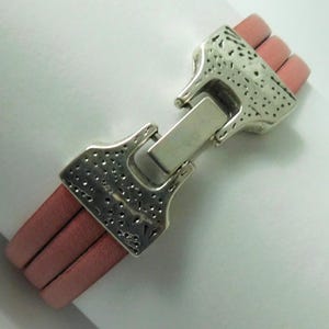 Puede incluir: Una pulsera con tres cordones de cuero rosa paralelos unidos por un cierre de metal plateado. El cierre tiene una superficie texturizada y picada con un mecanismo de bisagra rectangular central que asegura los dos extremos de la pulsera.