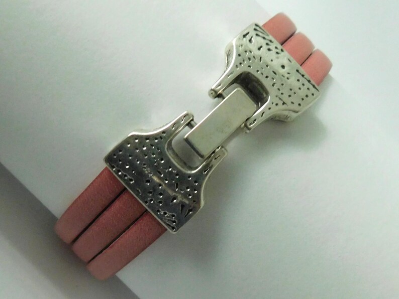 Puede incluir: Una pulsera con tres cordones de cuero rosa paralelos unidos por un cierre de metal plateado. El cierre tiene una superficie texturizada y picada con un mecanismo de bisagra rectangular central que asegura los dos extremos de la pulsera.