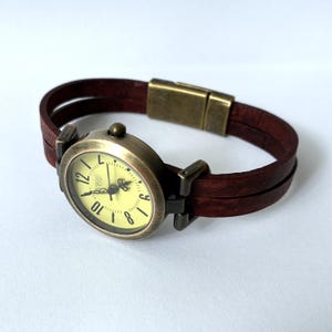 May include: A wristwatch featuring a round, antique-bronze-toned metal case with a pale yellow dial and black numerals. The watch has a double-strand dark brown leather strap secured by a matching antique-bronze-toned rectangular magnetic clasp.