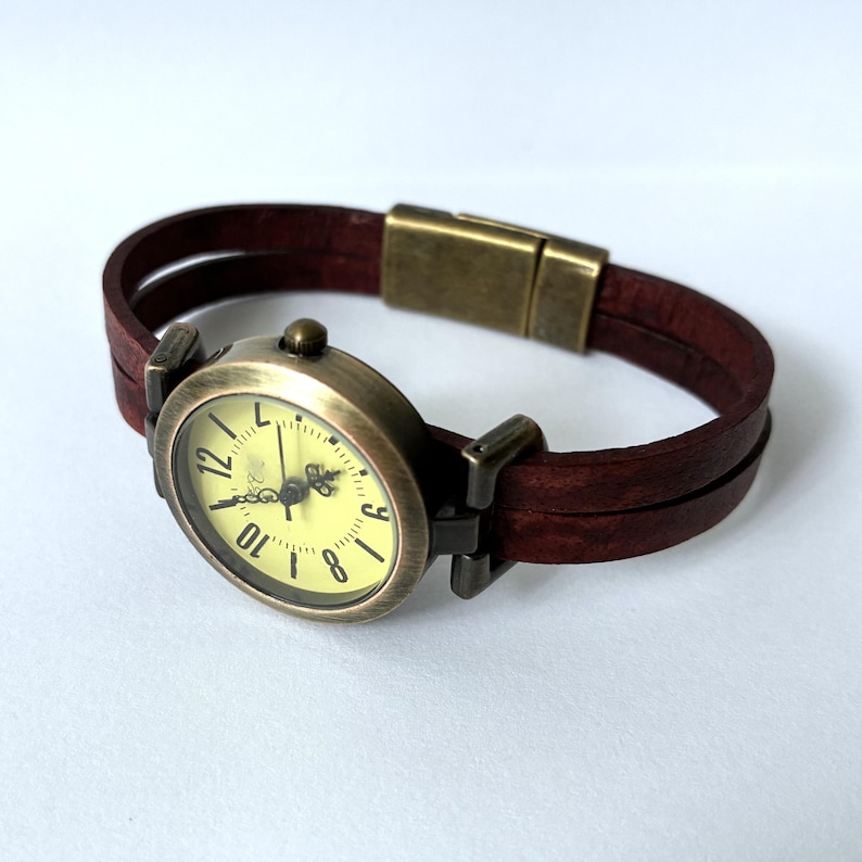 May include: A wristwatch featuring a round, antique-bronze-toned metal case with a pale yellow dial and black numerals. The watch has a double-strand dark brown leather strap secured by a matching antique-bronze-toned rectangular magnetic clasp.