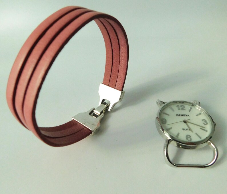 Puede incluir: Una pulsera de tres hilos en color rosa empolvado con un cierre de metal plateado y una esfera de reloj redonda plateada por separado con fondo blanco, que muestra el texto Geneva Elite, n&uacute;meros negros y manecillas plateadas, sin correa.