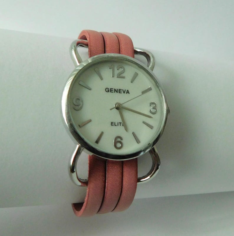 Puede incluir: Un reloj de pulsera con caja redonda plateada y esfera blanca con el texto GENEVA y ELITE. La esfera tiene marcadores de hora plateados y n&uacute;meros en las posiciones de las 3, 6, 9 y 12. El reloj cuenta con una correa de cuero rosa de varias tiras.