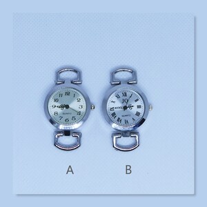 May include: Two silver wristwatches with white faces and black Roman numeral hour markers. The watch on the left has black Arabic numeral minute markers. The watch on the right has a black logo that says "JQ" and the word "QUARTZ" printed below it. Both watches have silver bands.