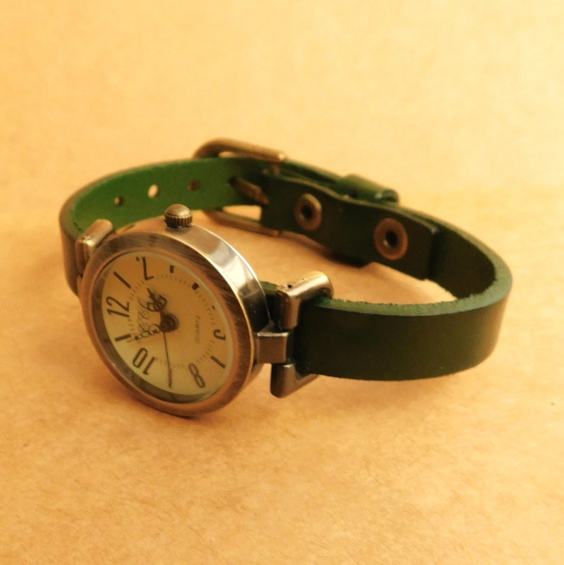 Op de afbeelding: Een ronde, zilverkleurige polshorloge met een witte wijzerplaat en zwarte Romeinse cijfers. Het horloge heeft een donkergroen leren bandje met een zilveren gesp.