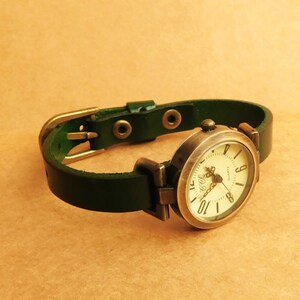 Op de afbeelding: Een groen leren polshorloge met een ronde, cr&egrave;mekleurige wijzerplaat en zwarte Romeinse cijfers. Het horloge heeft een gouden kast en gesp.