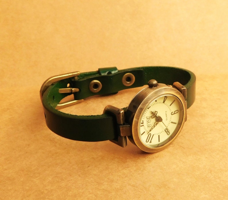 Op de afbeelding: Een groen leren polshorloge met een ronde, cr&egrave;mekleurige wijzerplaat en zwarte Romeinse cijfers. Het horloge heeft een gouden kast en gesp.