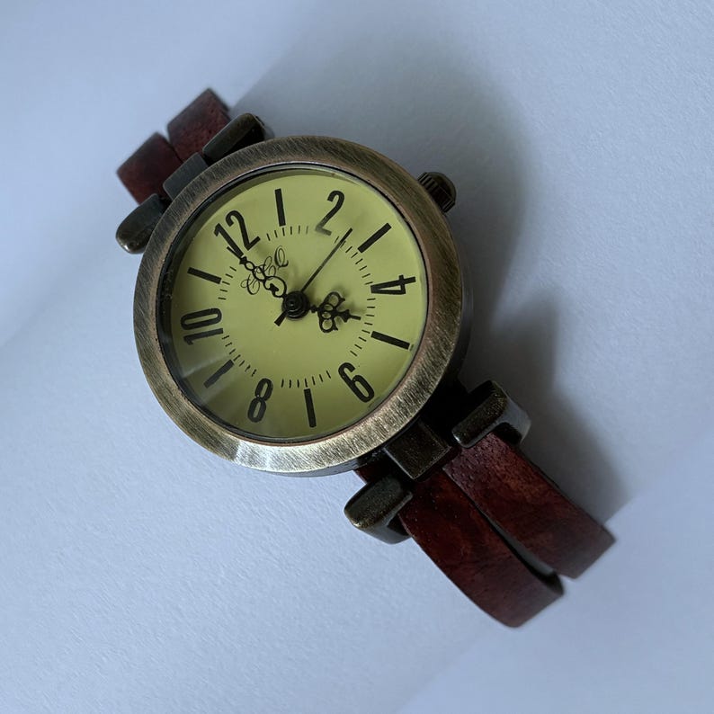 May include: A round analog wristwatch with a distressed bronze-tone metal case and a pale yellow face featuring black Arabic numerals and ornate hands. The watch is attached to a dark brown leather strap with a split-band design.