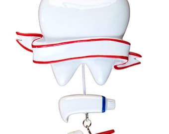 May include: A white tooth ornament with a red ribbon and a white banner. The tooth is hanging from a string with a white tube of toothpaste and a red toothbrush hanging below.